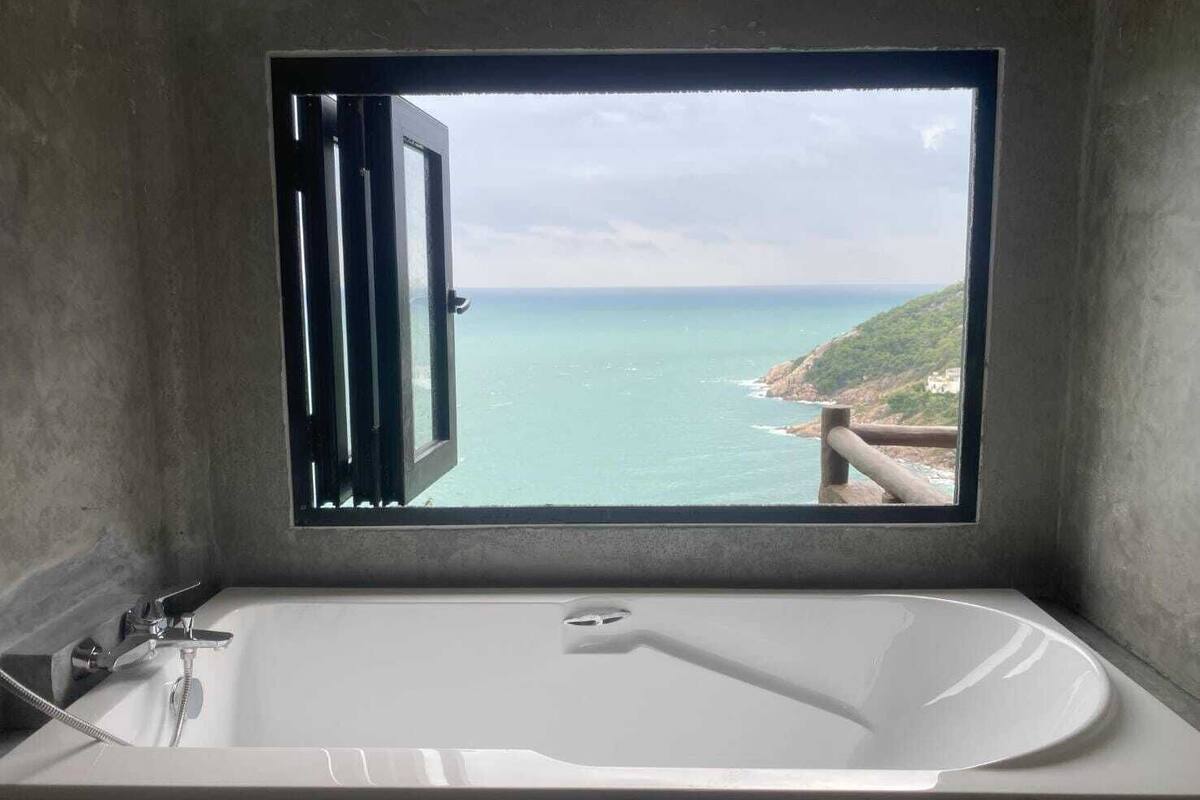 Un baño con bañera y ventana con vistas al mar y a la costa rocosa.