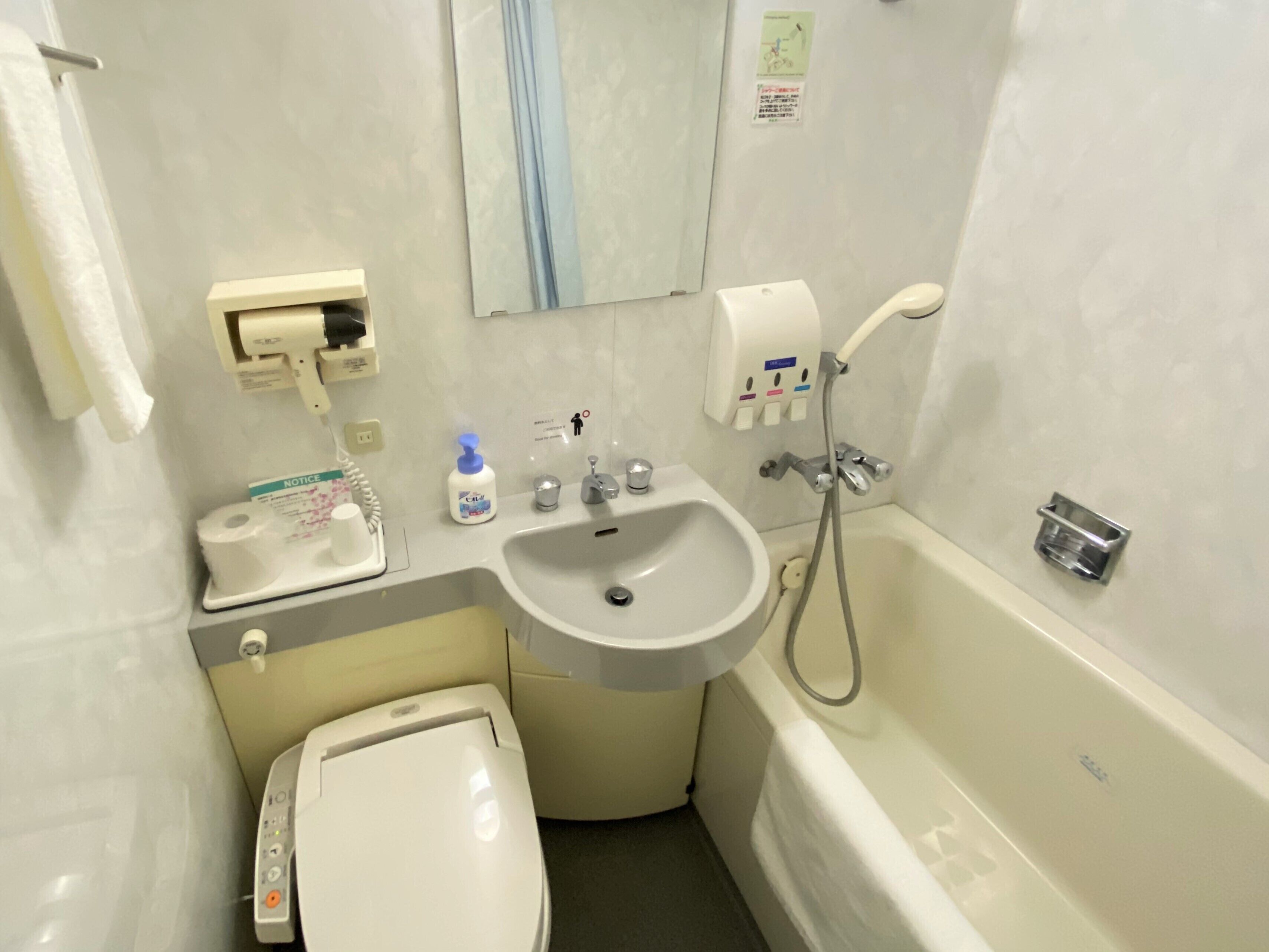 A small bathroom with a toilet, sink, and bathtub. There is a hand soap dispenser, a paper towel dispenser, and a toilet paper roll.