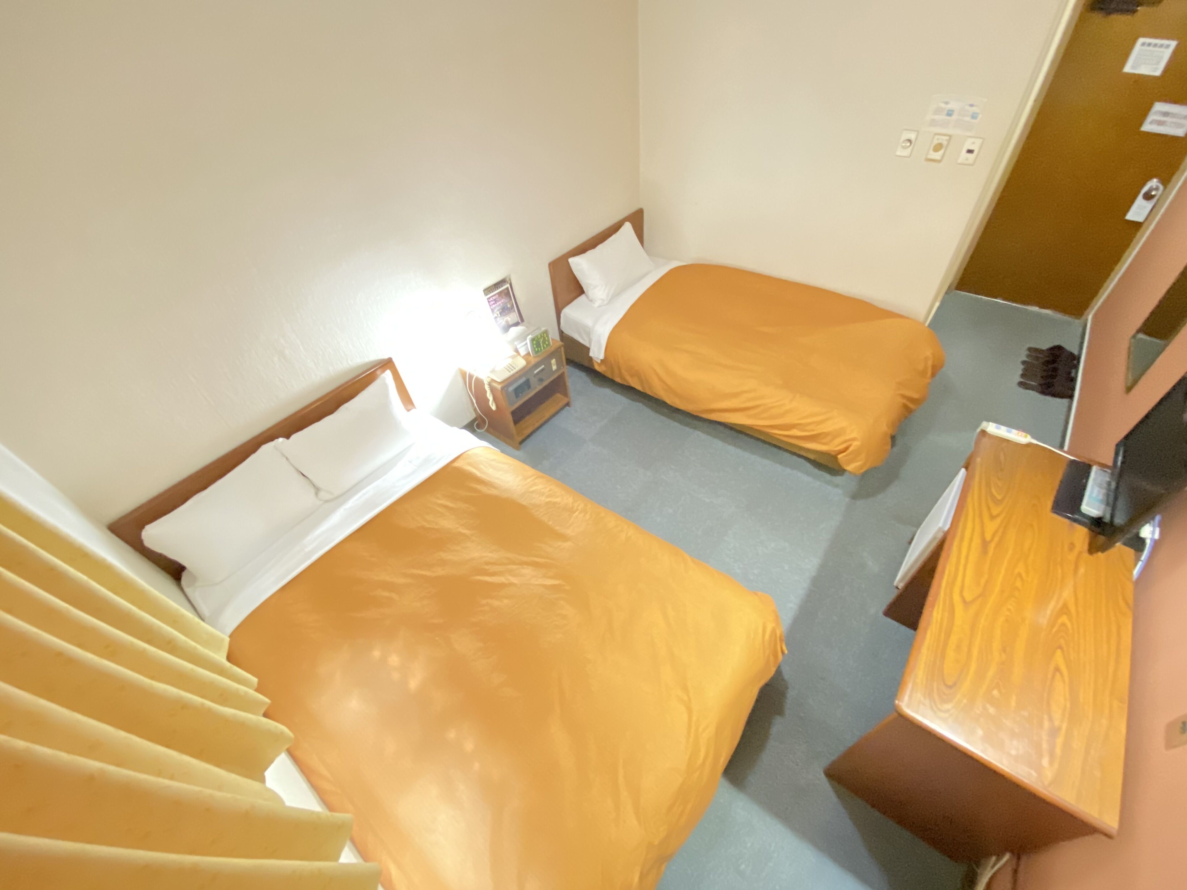 Habitación de hotel con dos camas, cada una con cabecero de madera, una mesita de noche pequeña y una mesita auxiliar. Se observa un escritorio de madera con una silla y un espejo.