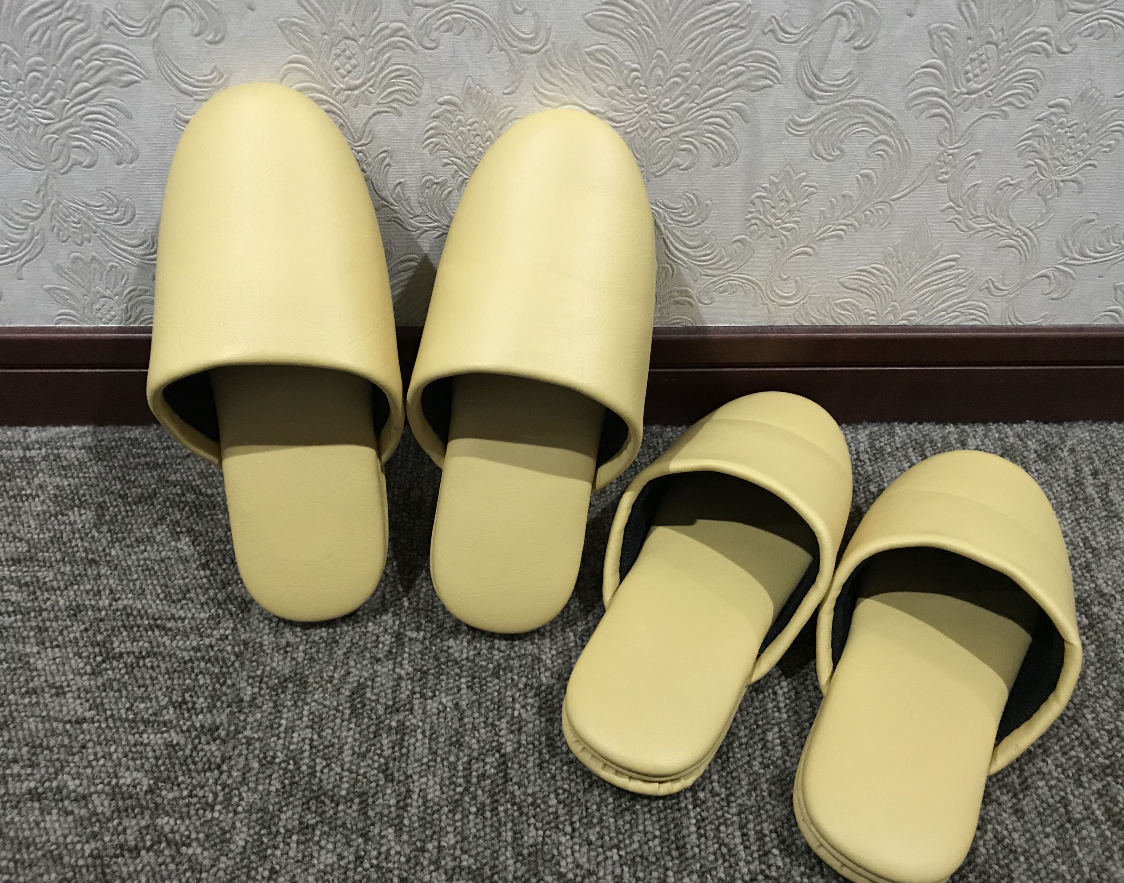 Une paire de pantoufles jaunes sur un tapis gris, avec un mur à motifs floraux en arrière-plan.