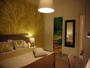 A hotel room with a bed, a nightstand, a lamp, a painting of a forest path, and a mirror. - In Roma Life (Rome)