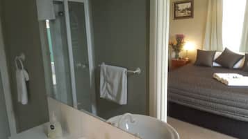 Ein Badezimmer mit einer gläsernen Duschkabine, einer weißen Badewanne und einem Waschbecken mit einer Flasche Pure Seife. Der Raum verfügt über einen großen Spiegel und ein Bett mit dunkler Bettdecke und Kissen.