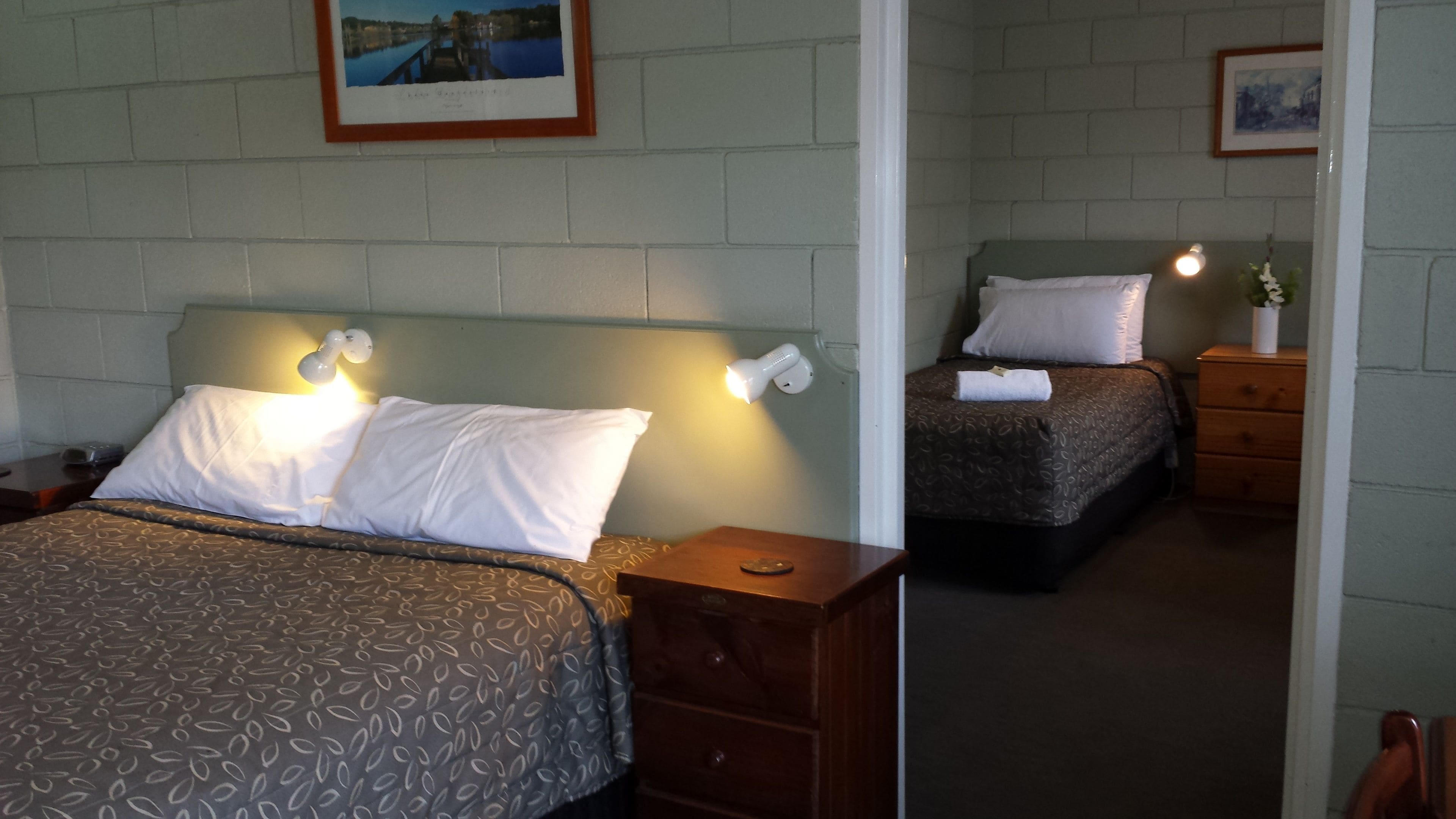 A hotel room with two beds, each with a headboard and bedside lamps. There is a wooden dresser with a mirror and a small table with a lamp.