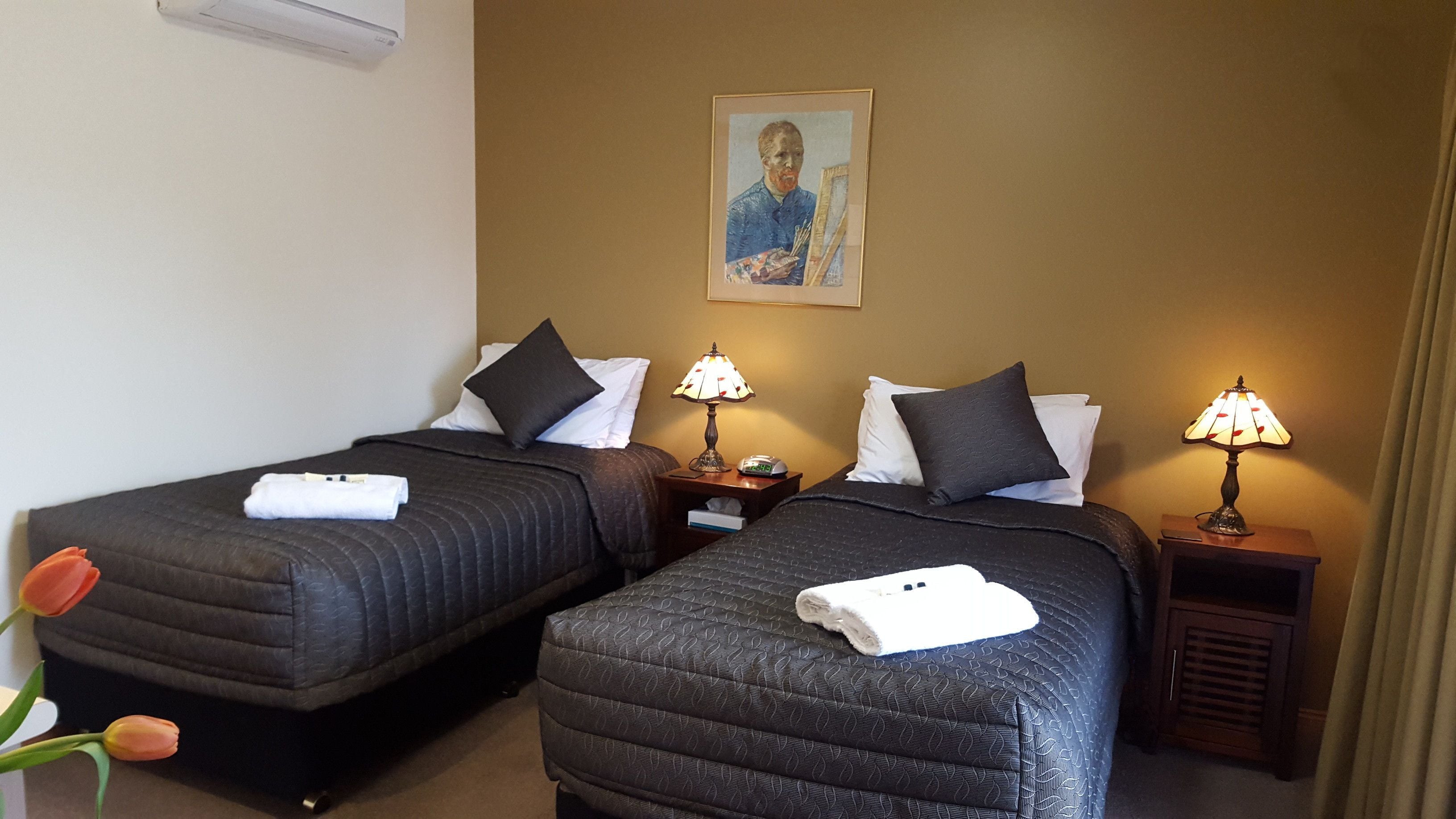 Une chambre d’hôtel avec deux lits, chacun recouvert d’une courtepointe matelassée de couleur sombre, agrémentée d’oreillers blancs et d’une petite table de chevet avec une lampe. Un tableau encadré est accroché au mur au-dessus des lits.