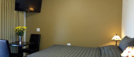 A hotel room with a bed, a desk, a chair, a TV mounted on the wall, a lamp, and a vase with flowers.
