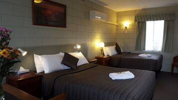 A hotel room with two beds, each with a headboard, bedside lamps, and a bedside table with a tissue box. There is a window with curtains, a wall-mounted air conditioner, and a framed picture above the beds.