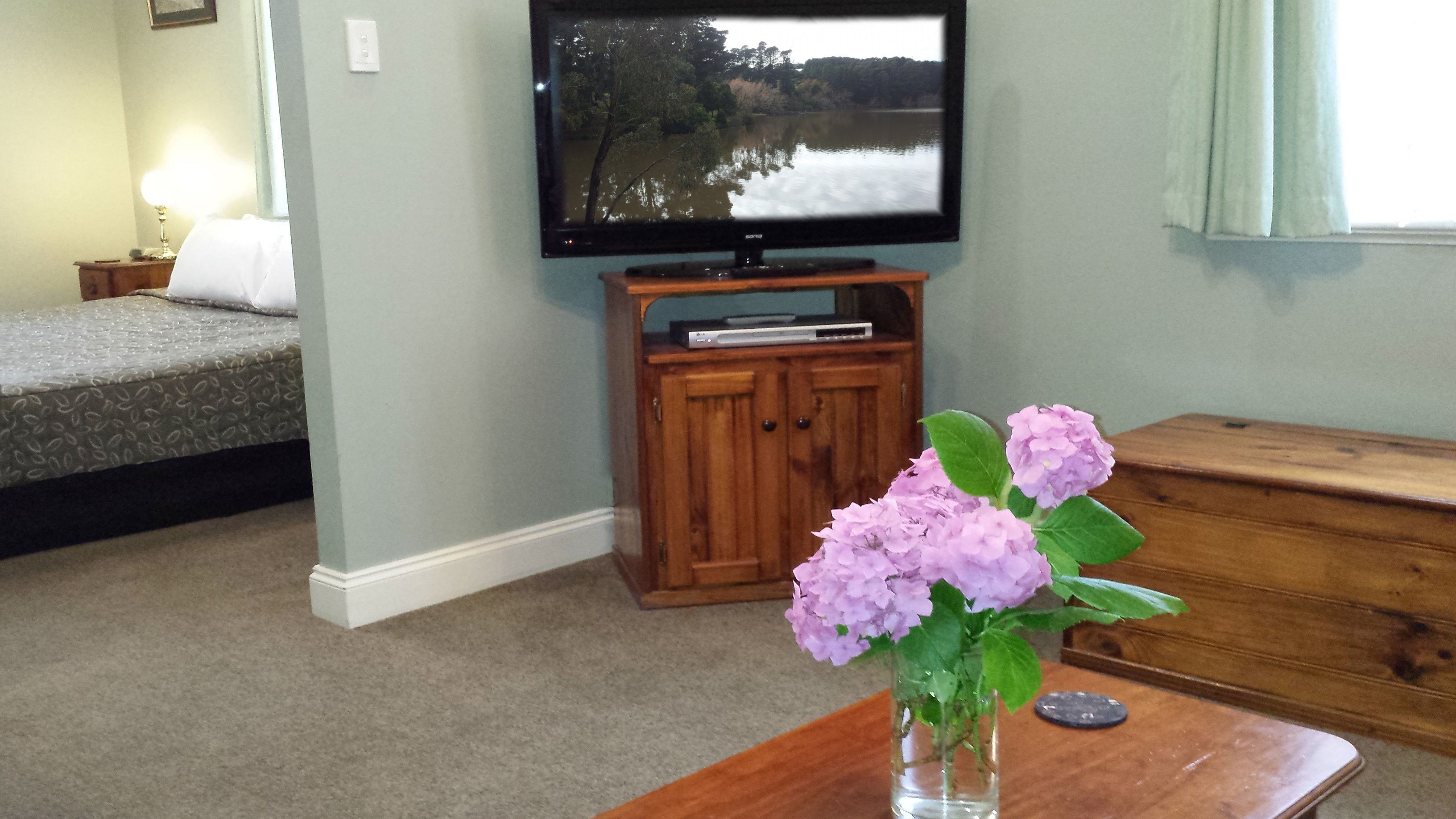 A bedroom with a bed, a television on a wooden stand, and a vase with flowers on a table.