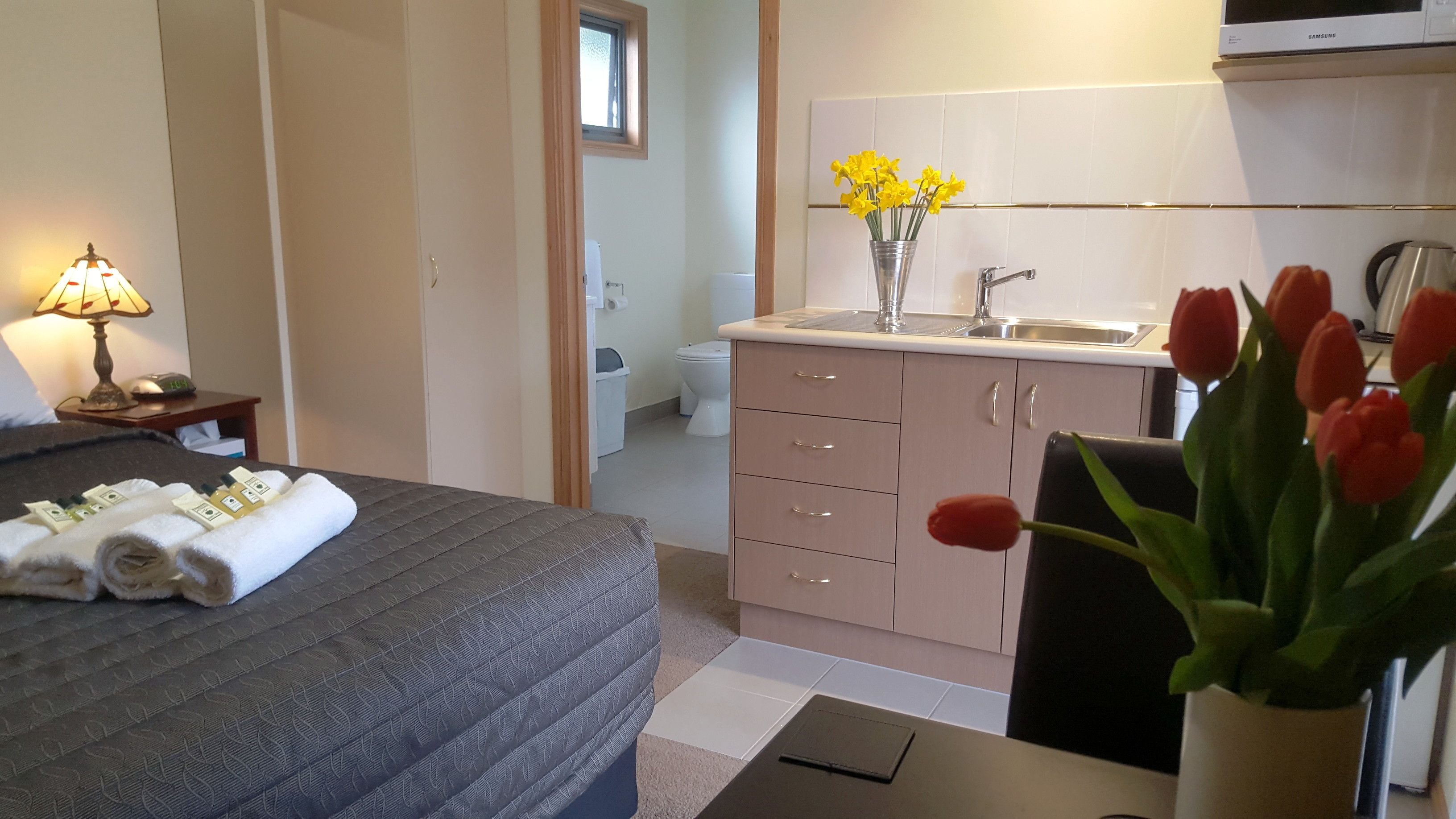 Een hotelkamer met een bed, bureau, stoel, bureaulamp, vaas met bloemen, wastafel en toilet.
