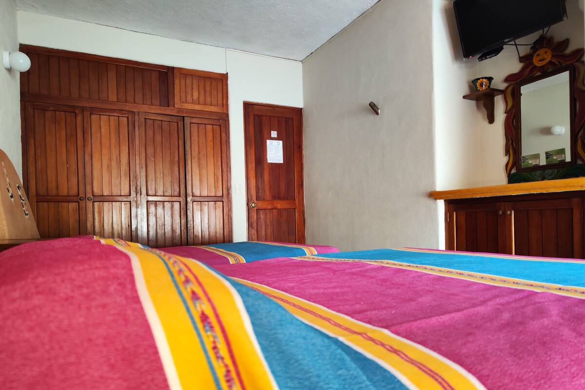 Ein Zimmer mit einem Bett, das mit farbenfroher Bettwäsche bezogen ist, Holzschränken, einem Fernseher und einem Spiegel mit dekorativem Rahmen.