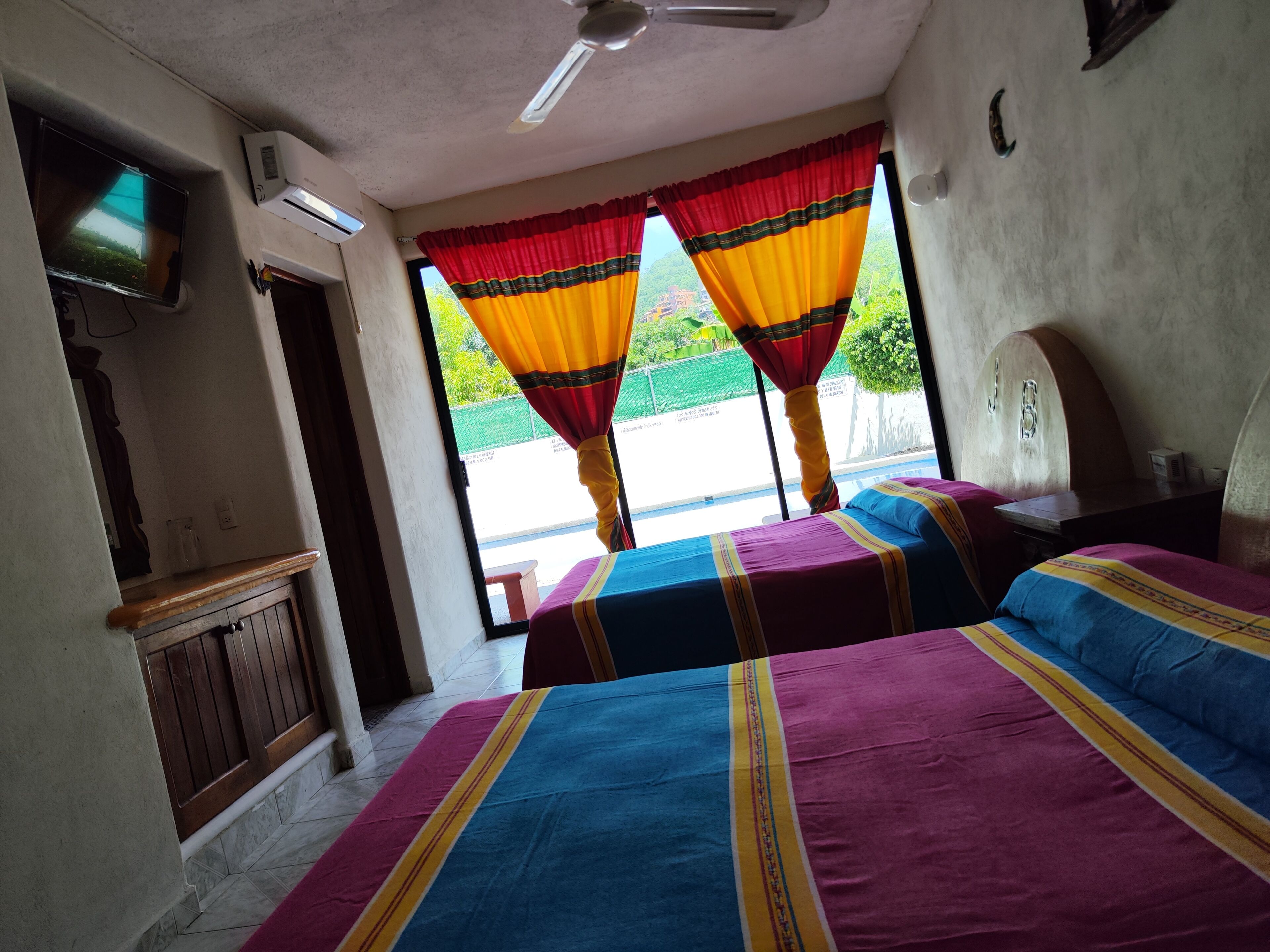 A room with two beds, a ceiling fan, a TV, and a window with curtains.