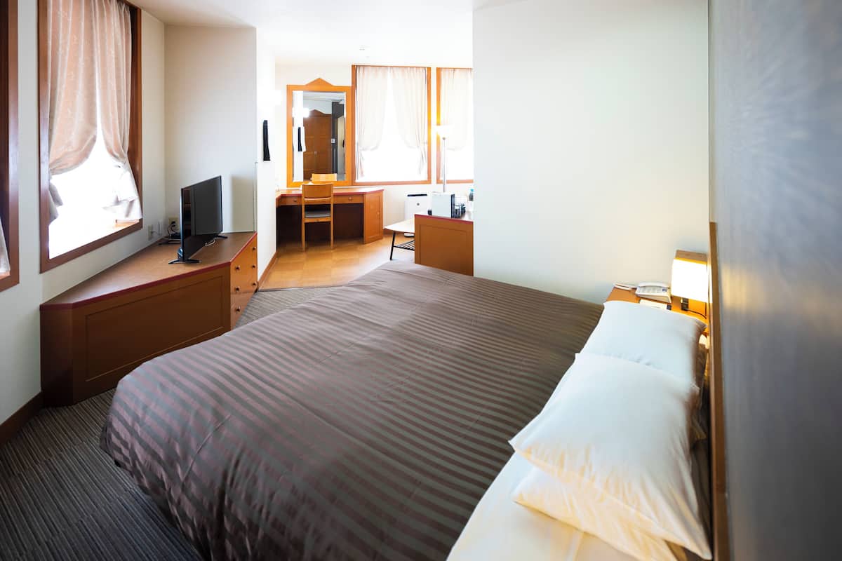 Une chambre d’hôtel avec un grand lit, un bureau avec un ordinateur, une chaise, un miroir et une fenêtre avec des rideaux.