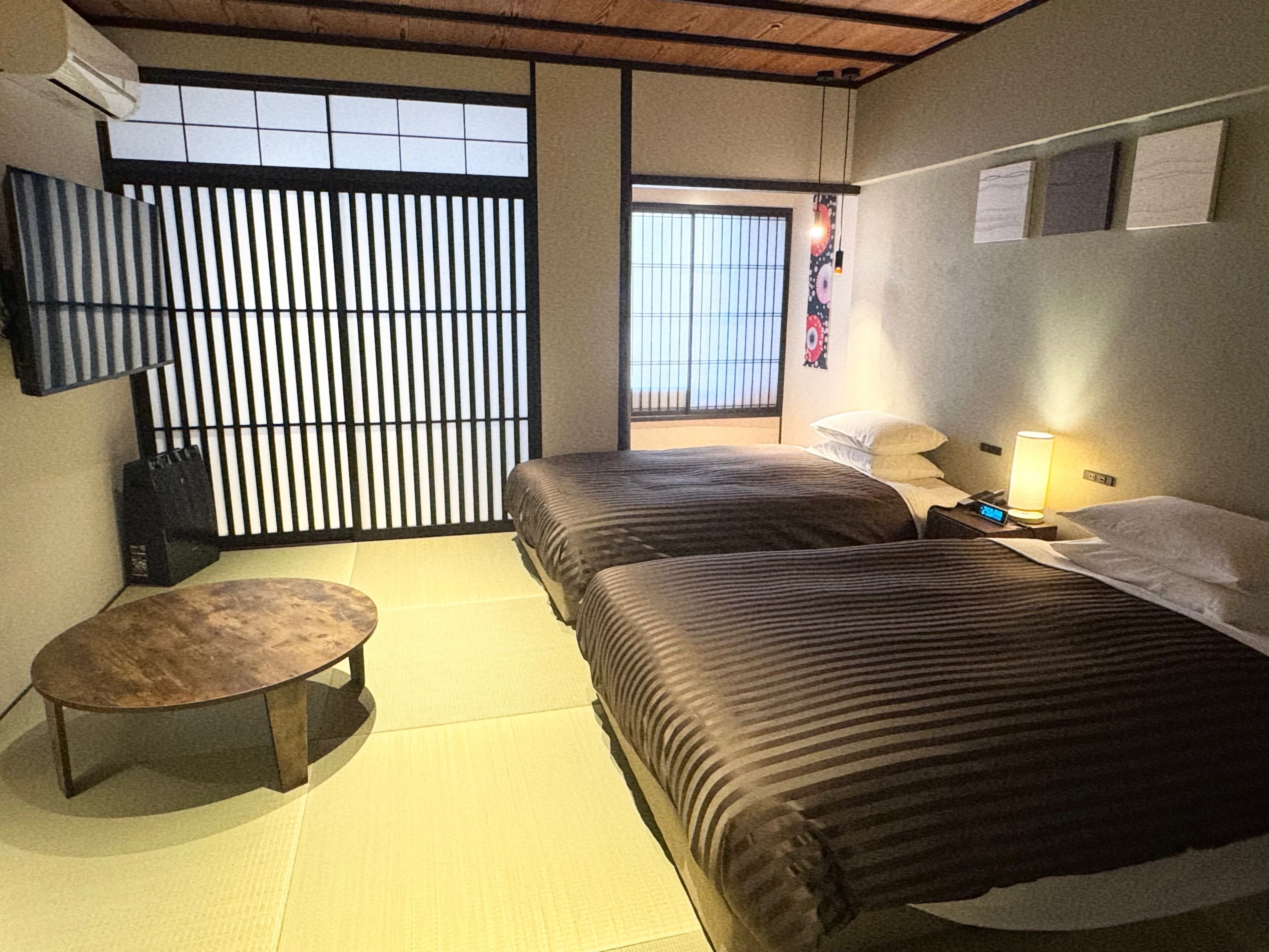 A hotel room with two beds, a small table, a wall-mounted TV, and a sliding door to a balcony.