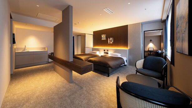 A modern hotel room with a bed, bedside table, chair, and a desk with a lamp.