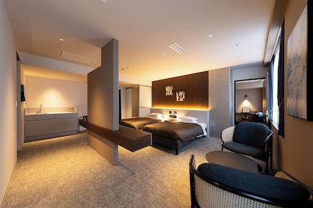 A modern hotel room with a bed, bedside table, chair, and a desk with a lamp.