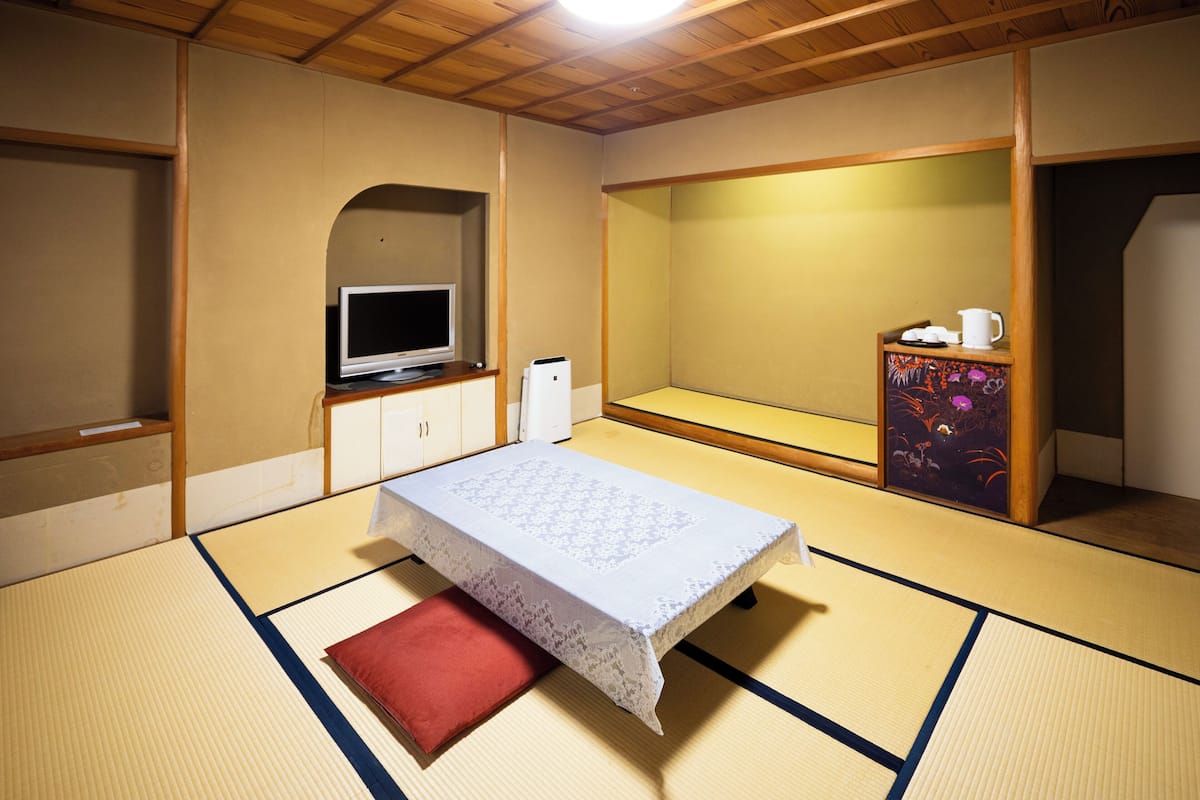 Une pièce de style japonais traditionnel, avec un sol recouvert de tatamis, un lit bas agrémenté d’un coussin rouge, un téléviseur posé sur un meuble et un petit meuble décoré de motifs floraux.
