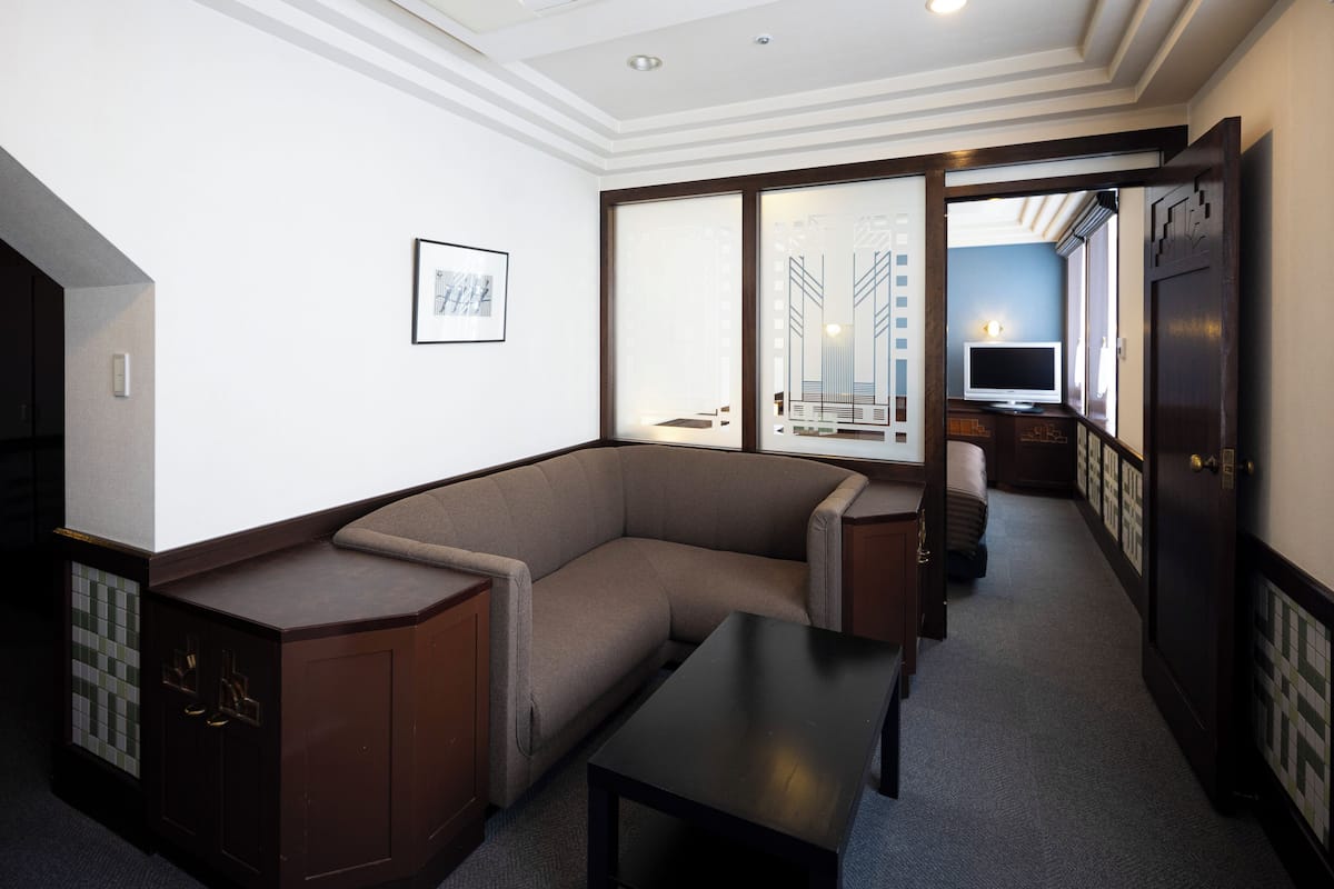 Une chambre d’hôtel moderne, équipée d’un canapé d’angle, d’une petite table, d’un meuble, d’une télévision et d’une porte donnant sur une autre pièce.