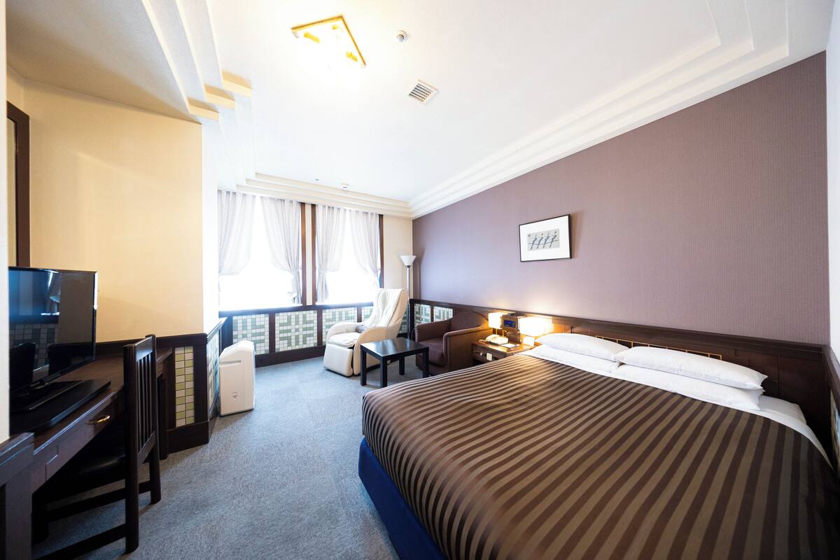 Une chambre d’hôtel avec un lit, un bureau, une chaise, une table de chevet et une fenêtre avec des rideaux.