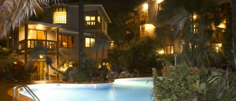 A nighttime view of a hotel with a swimming pool, palm trees, and well-lit buildings.