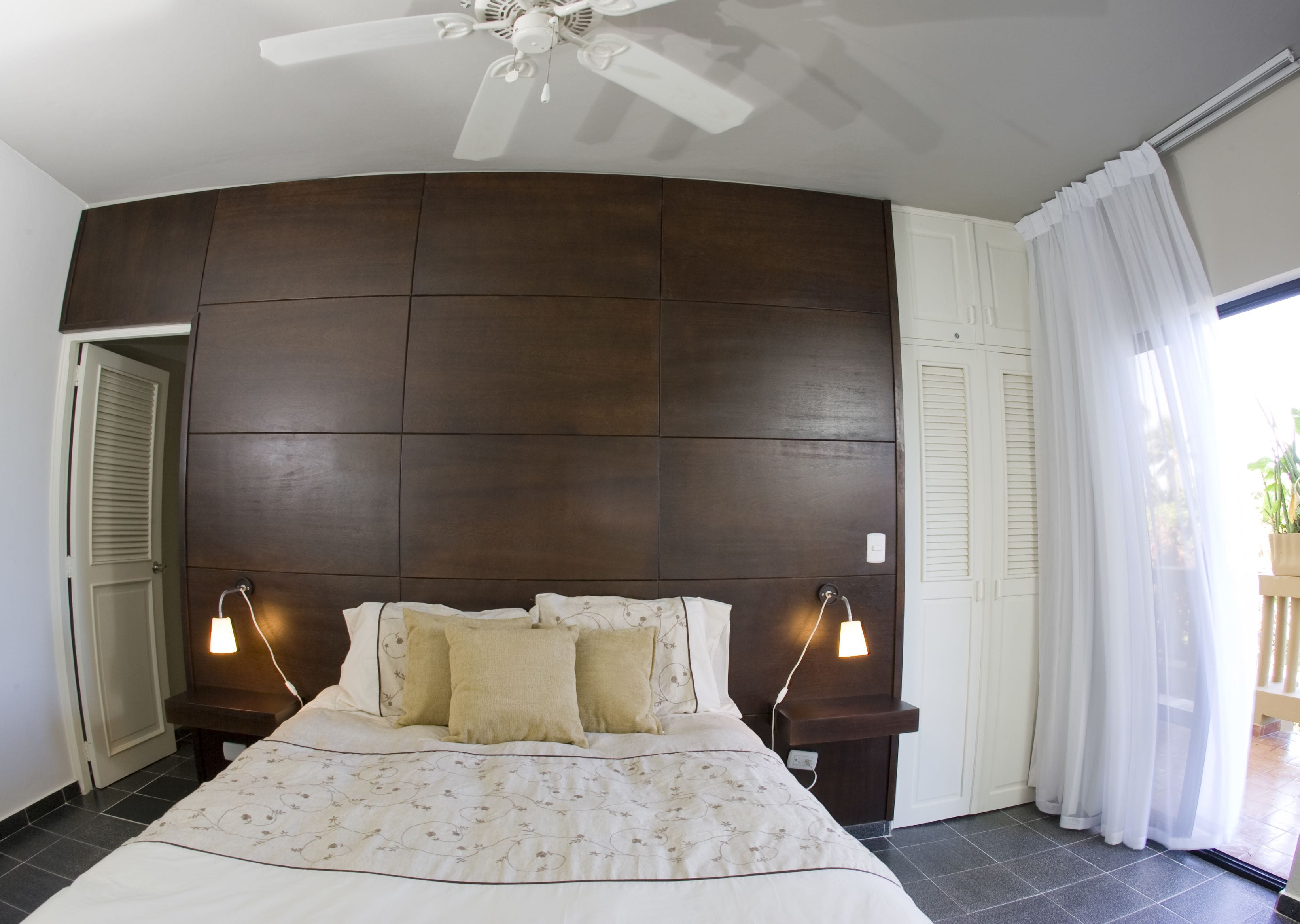 A bedroom with a large bed, two bedside lamps, a ceiling fan, and a view of the outdoors through a window with sheer curtains.