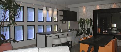 A modern kitchen with a central island, a dining area with a black table and orange chairs, and a seating area with a white sofa.