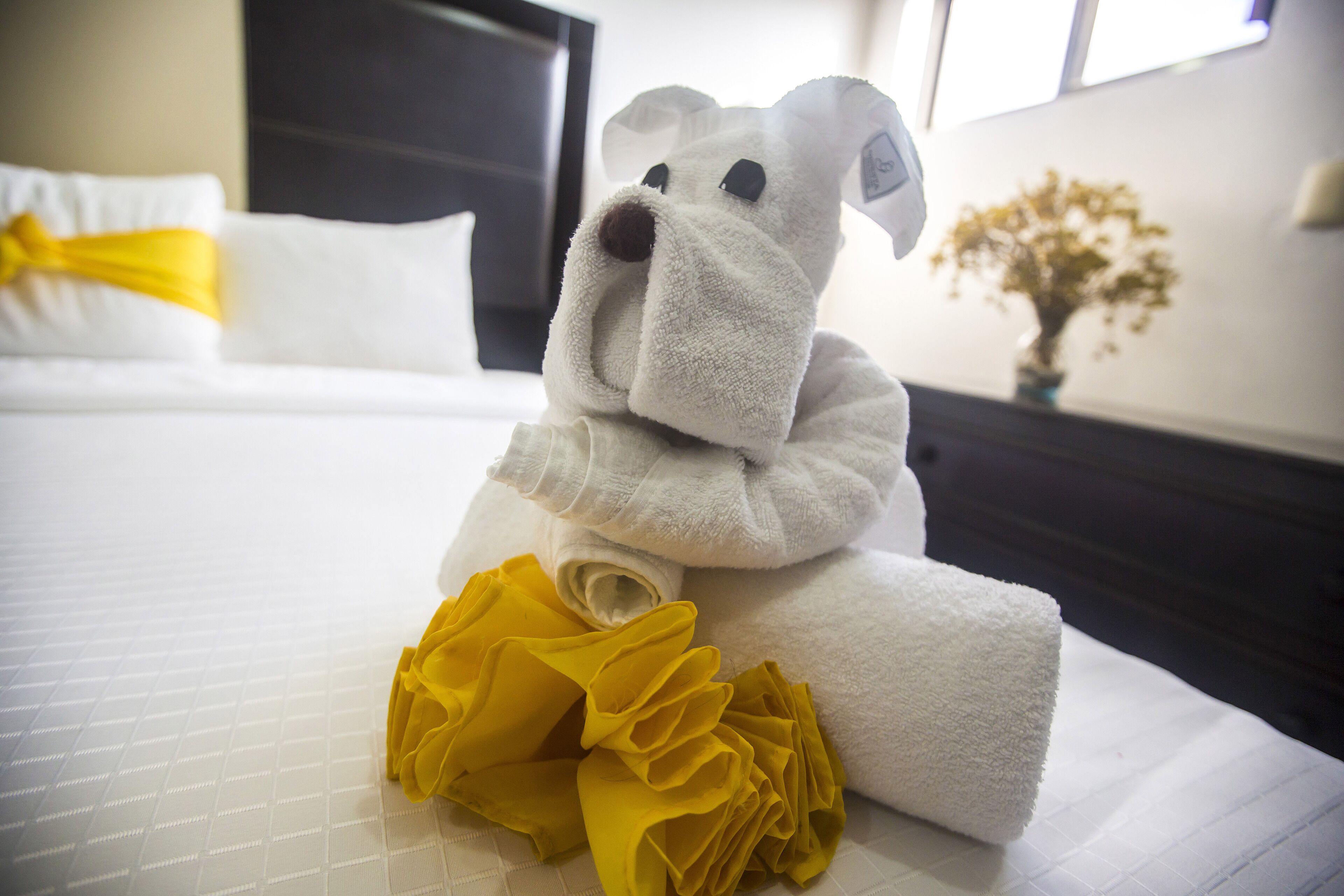 Una cama bien hecha con una almohada blanca y un lazo amarillo, una toalla blanca con un lazo amarillo, y un peluche sobre la cama.