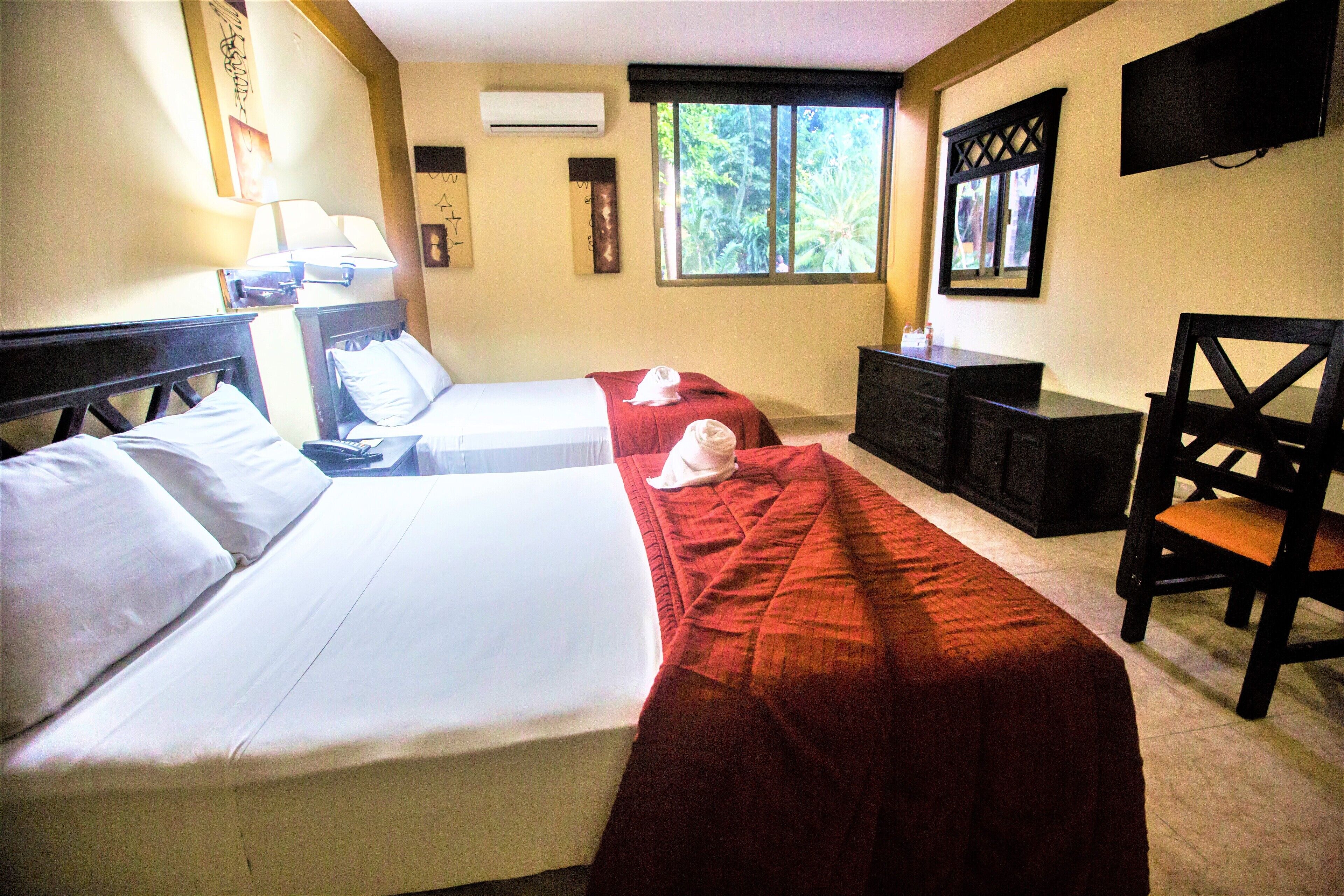 Habitación de hotel con dos camas, un escritorio, una silla, un televisor y una ventana con vistas a la vegetación.