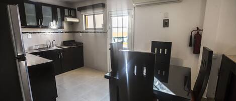 Cocina compacta con refrigerador, estufa, lavabo y gabinetes negros. Hay una ventana con vistas al exterior, y un equipo de aire acondicionado montado en la pared.
