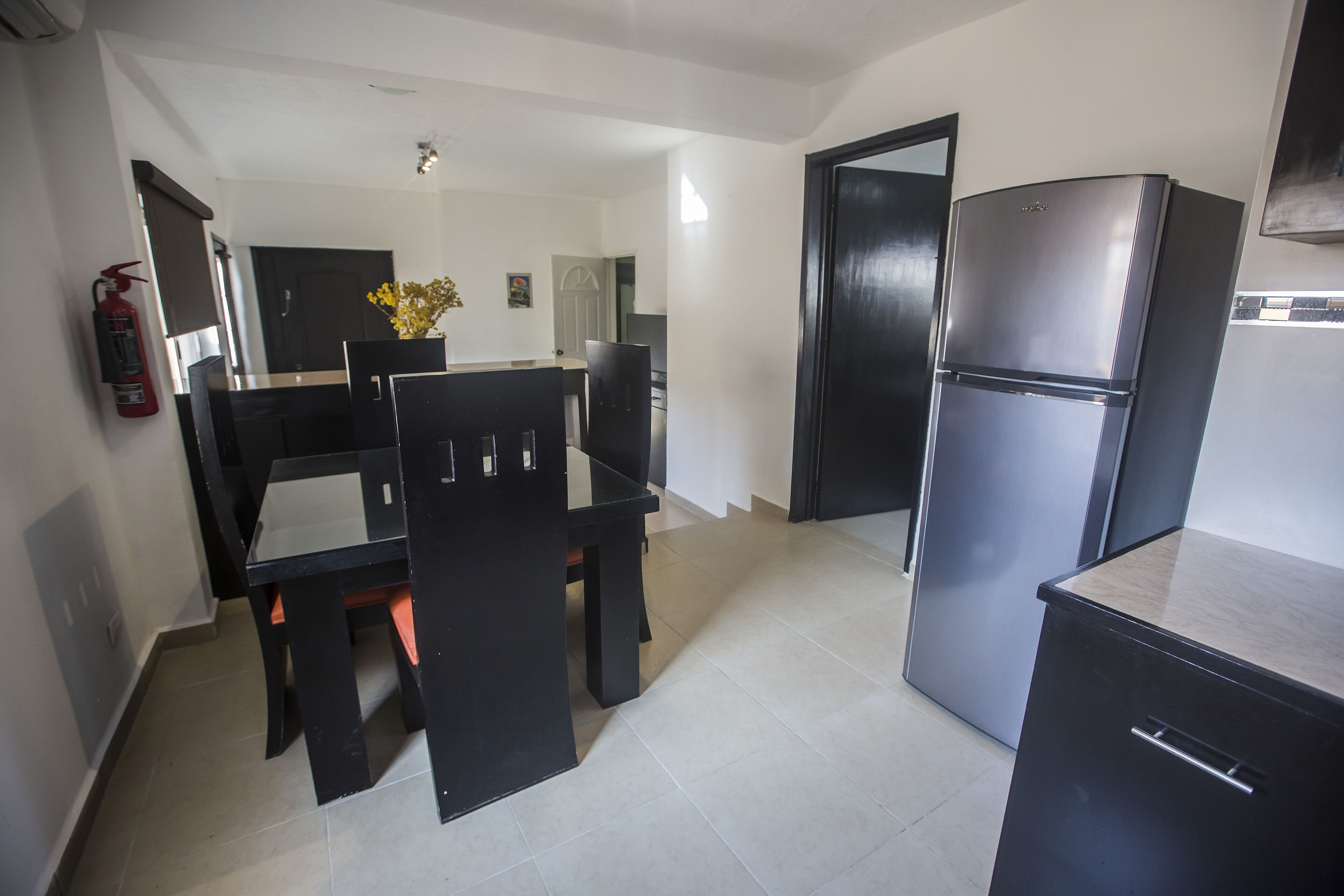 A compact kitchen with a dining table, chairs, a refrigerator, and a fire extinguisher.