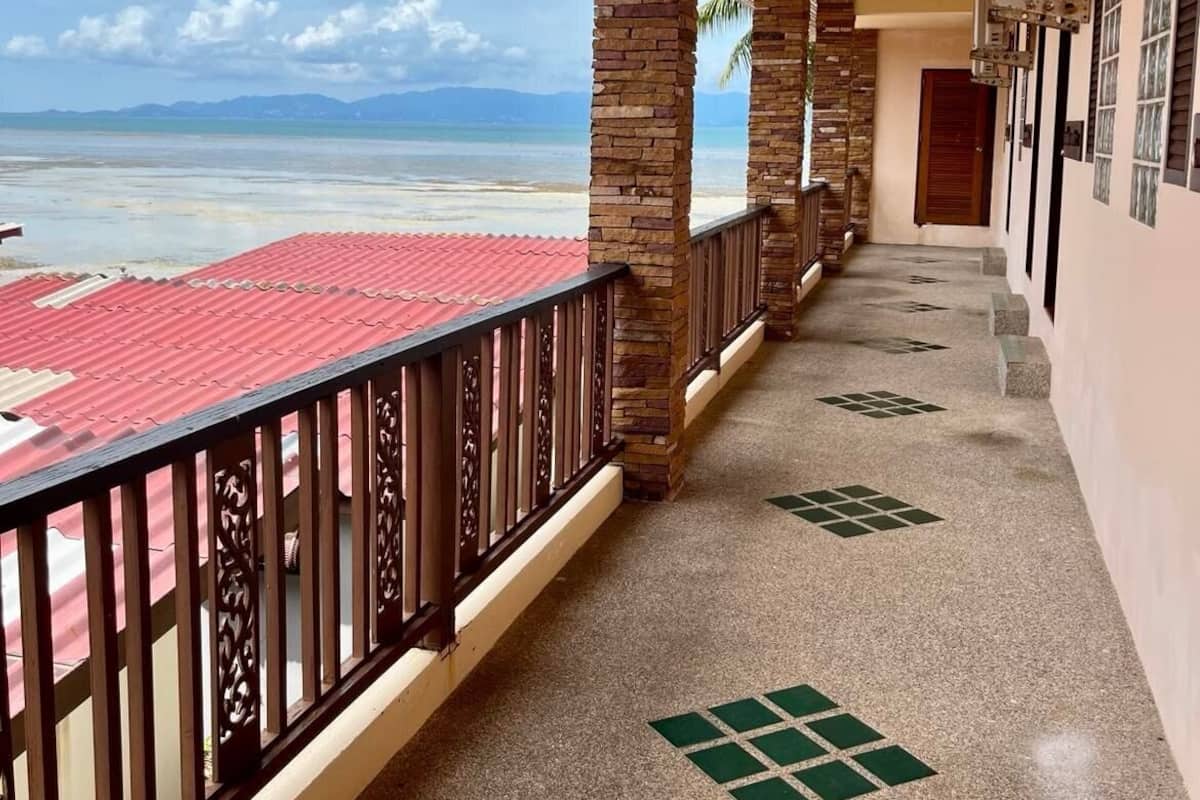 A corridor with a view of the beach, featuring a red-roofed balcony, stone pillars, and a tiled floor with green patterns.