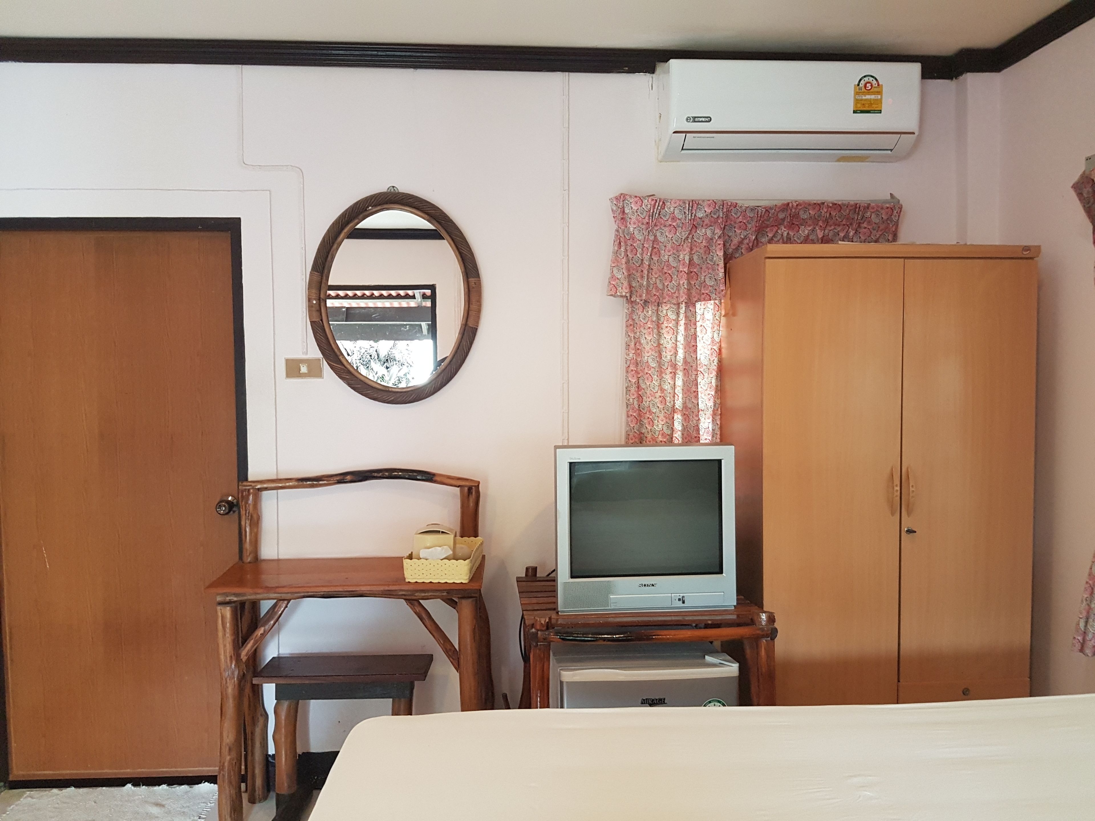 Une chambre avec une armoire en bois, un téléviseur posé sur un meuble en bois, un miroir et un climatiseur fixé au mur.