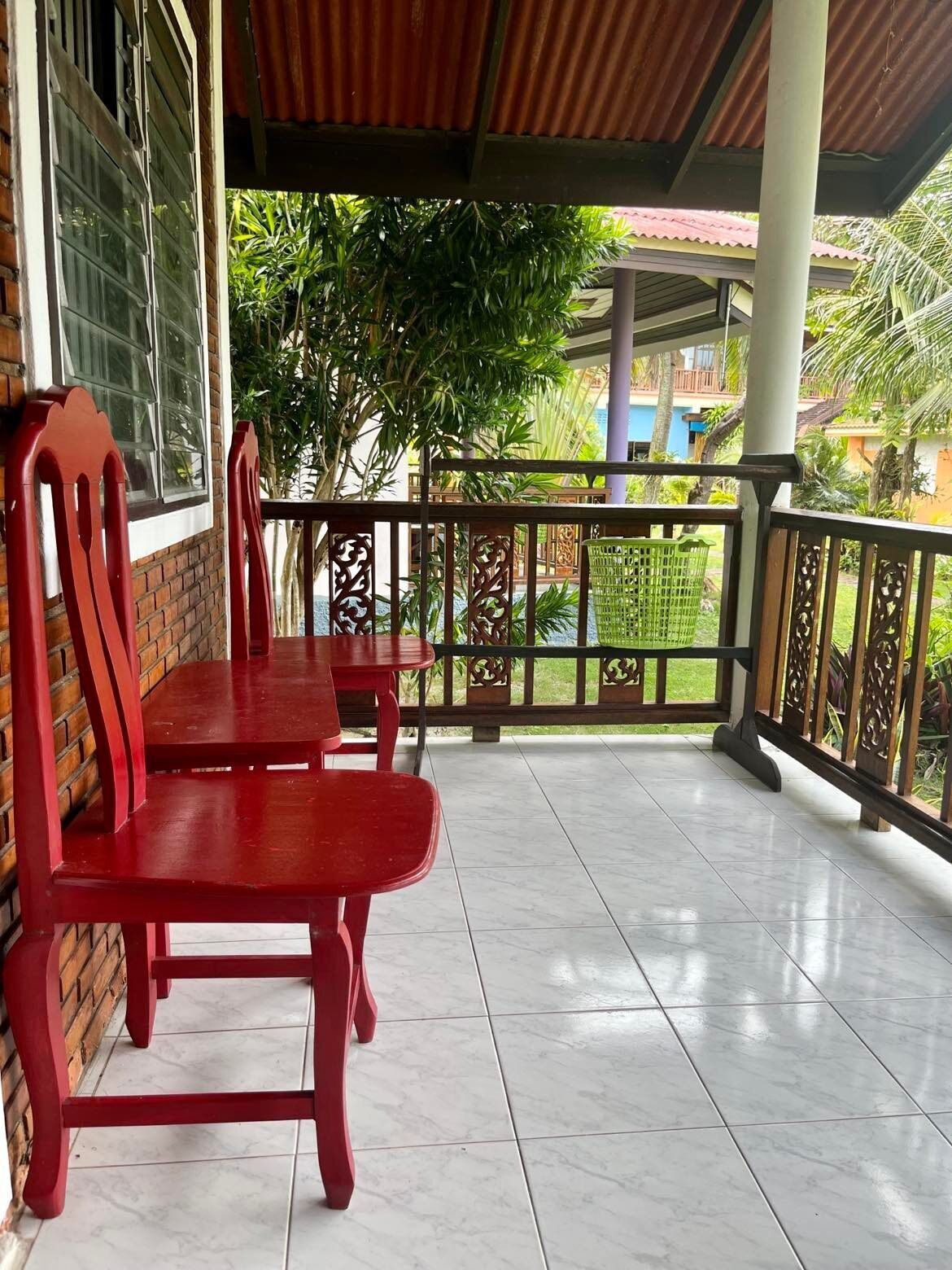 Een overdekt terras met twee rode houten stoelen en een tafel, een betegelde vloer, en zicht op groen en gebouwen op de achtergrond.