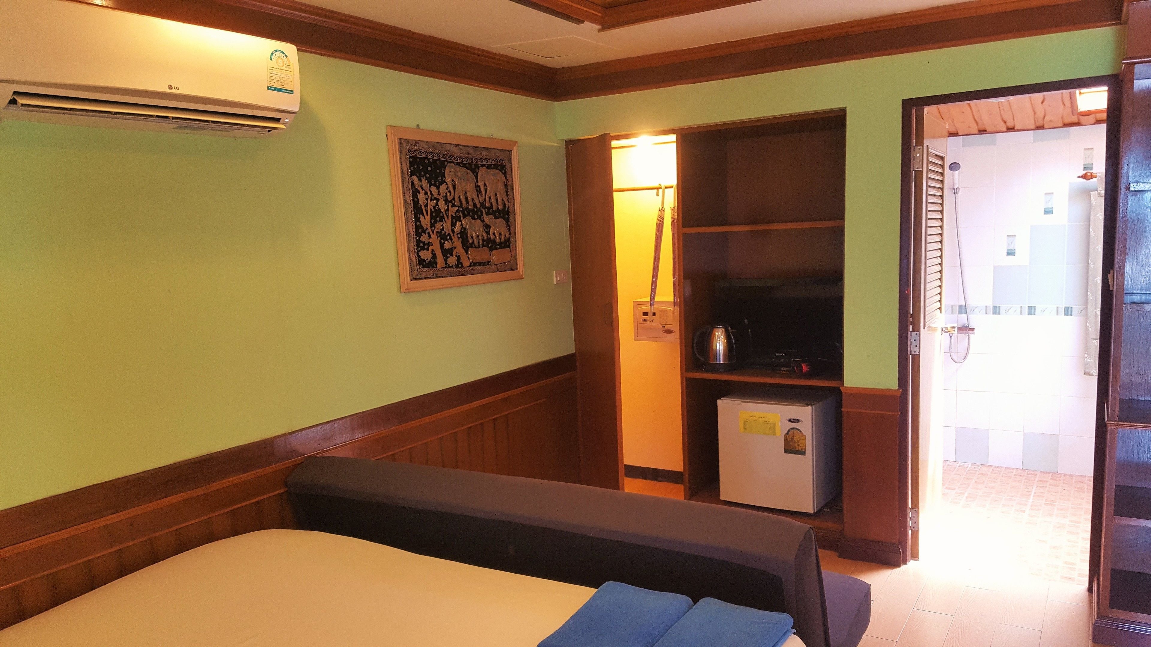 A hotel room with a bed, an air conditioner, a framed picture, a small refrigerator, and a bathroom visible through an open door.