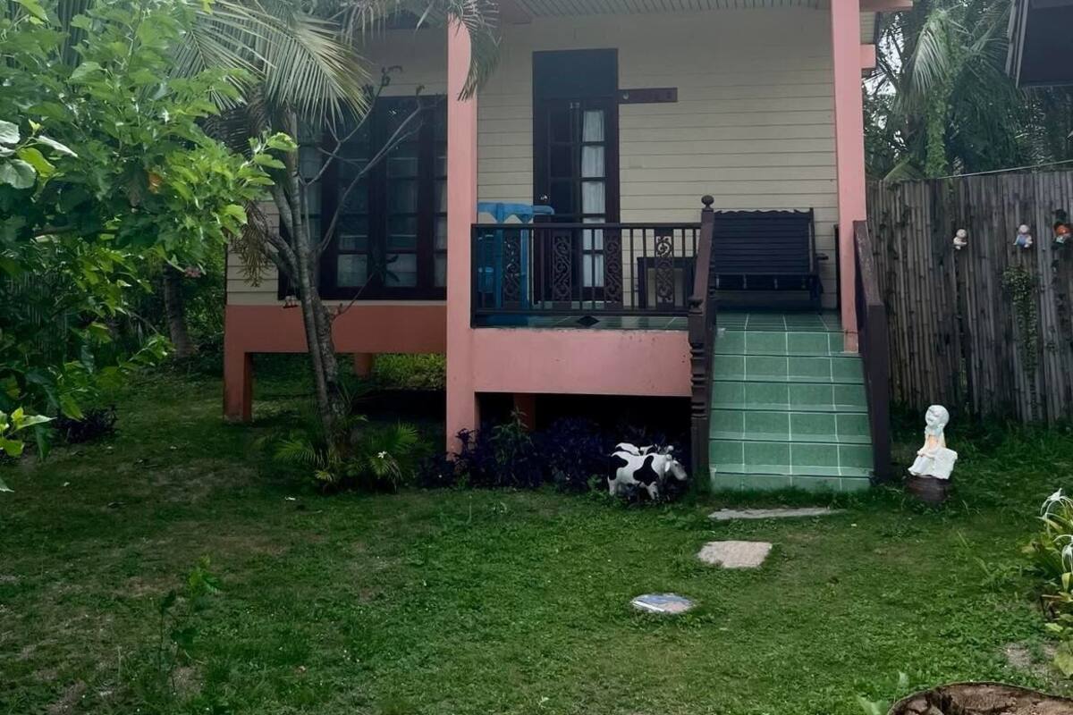 A small, single-story house with a pink exterior and a green roof. It features a front porch with a black railing and a small staircase leading up to the entrance. The surrounding area is lush with greenery, including palm trees and various plants.