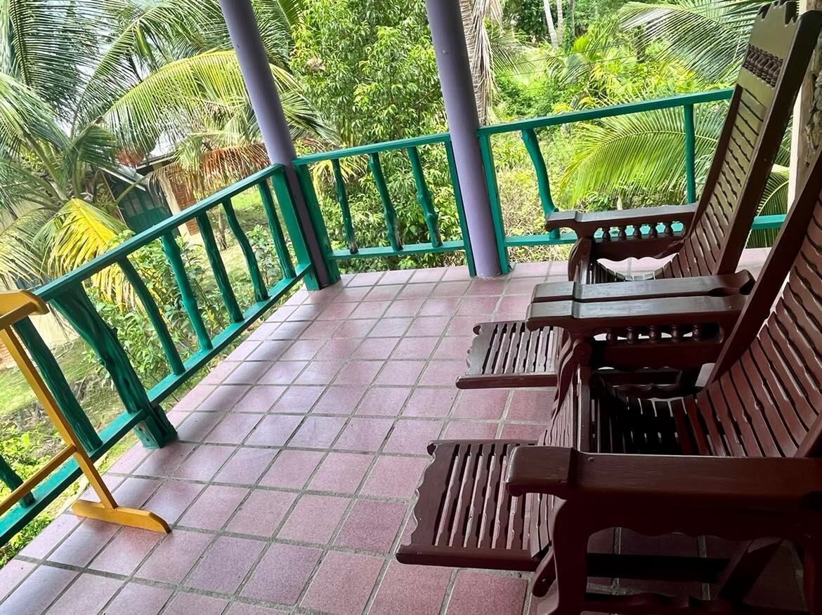 A covered outdoor seating area with benches and a tiled floor, surrounded by a green railing and lush greenery.