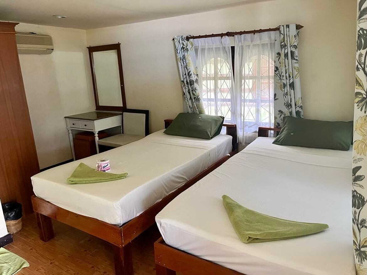 A room with two single beds, a desk with a chair, a mirror, and a window with floral curtains.