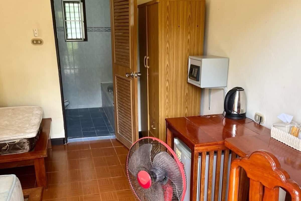 A compact room with a wooden table, a red standing fan, a wooden chair, a bed, and a wardrobe.