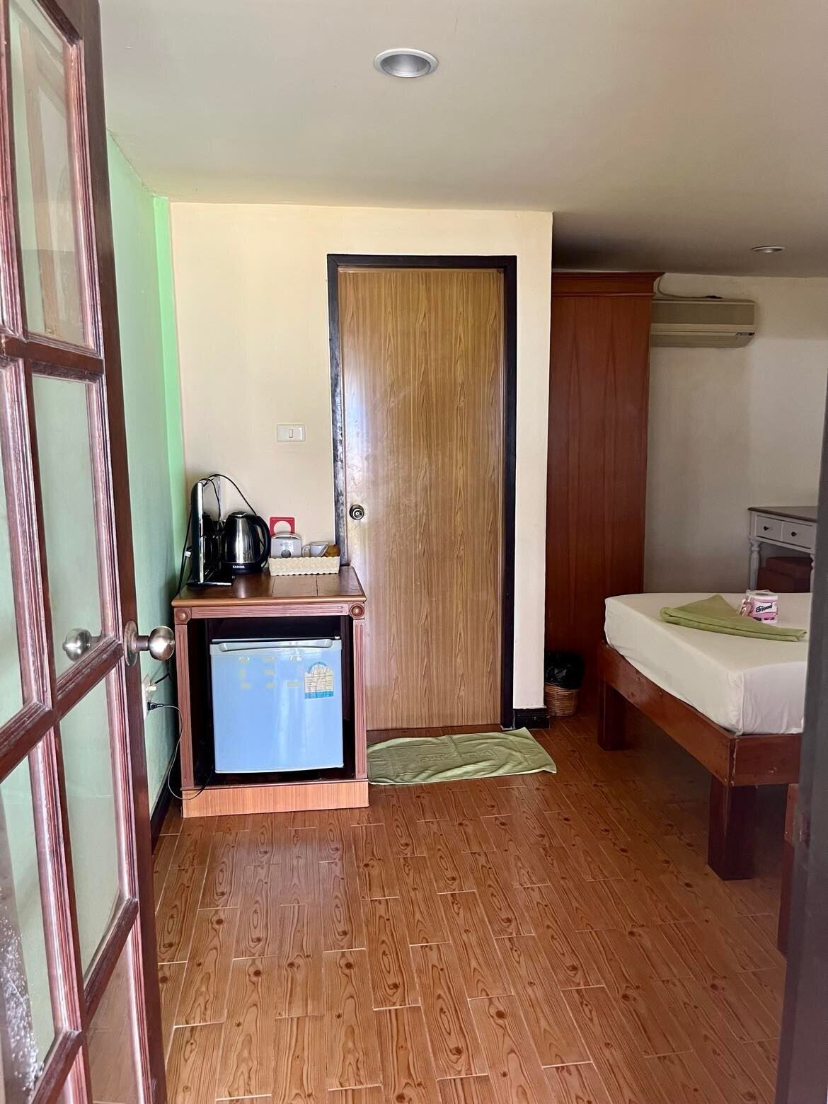 Una habitación compacta con piso de madera, una kitchenette pequeña con refrigerador, microondas y cafetera. Hay una cama con una manta verde y una mesita de noche con lámpara.