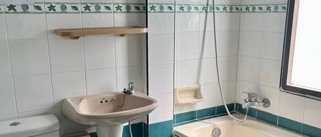 Un baño con inodoro, lavamanos y bañera. Hay un espejo sobre el lavamanos y un cabezal de ducha sobre la bañera. Las paredes están revestidas con un diseño azul y blanco.