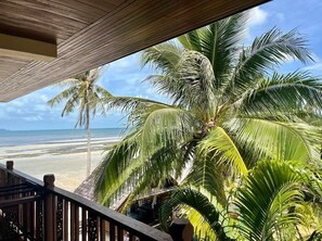 Un balcon en bois donnant sur une plage bordée de palmiers, avec un ciel dégagé.