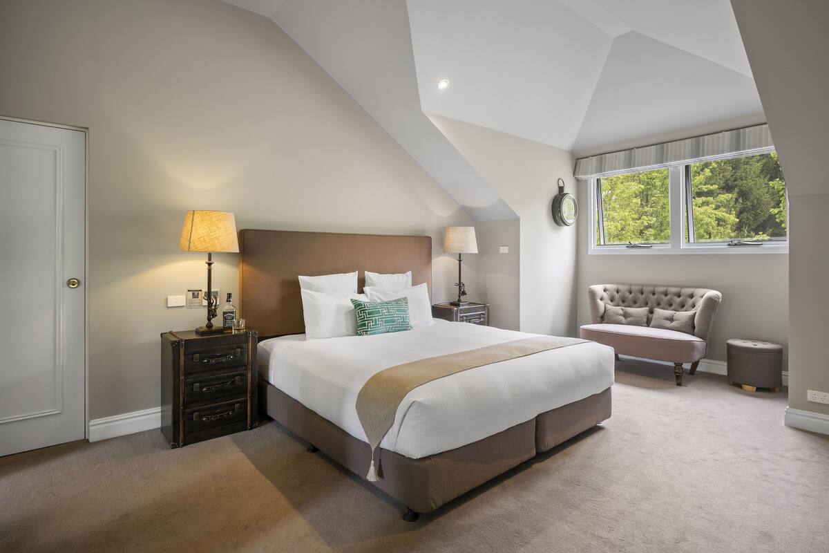 A bedroom with a large bed, two bedside tables, a nightstand, a lamp, a clock, a window with a view, and a chair.