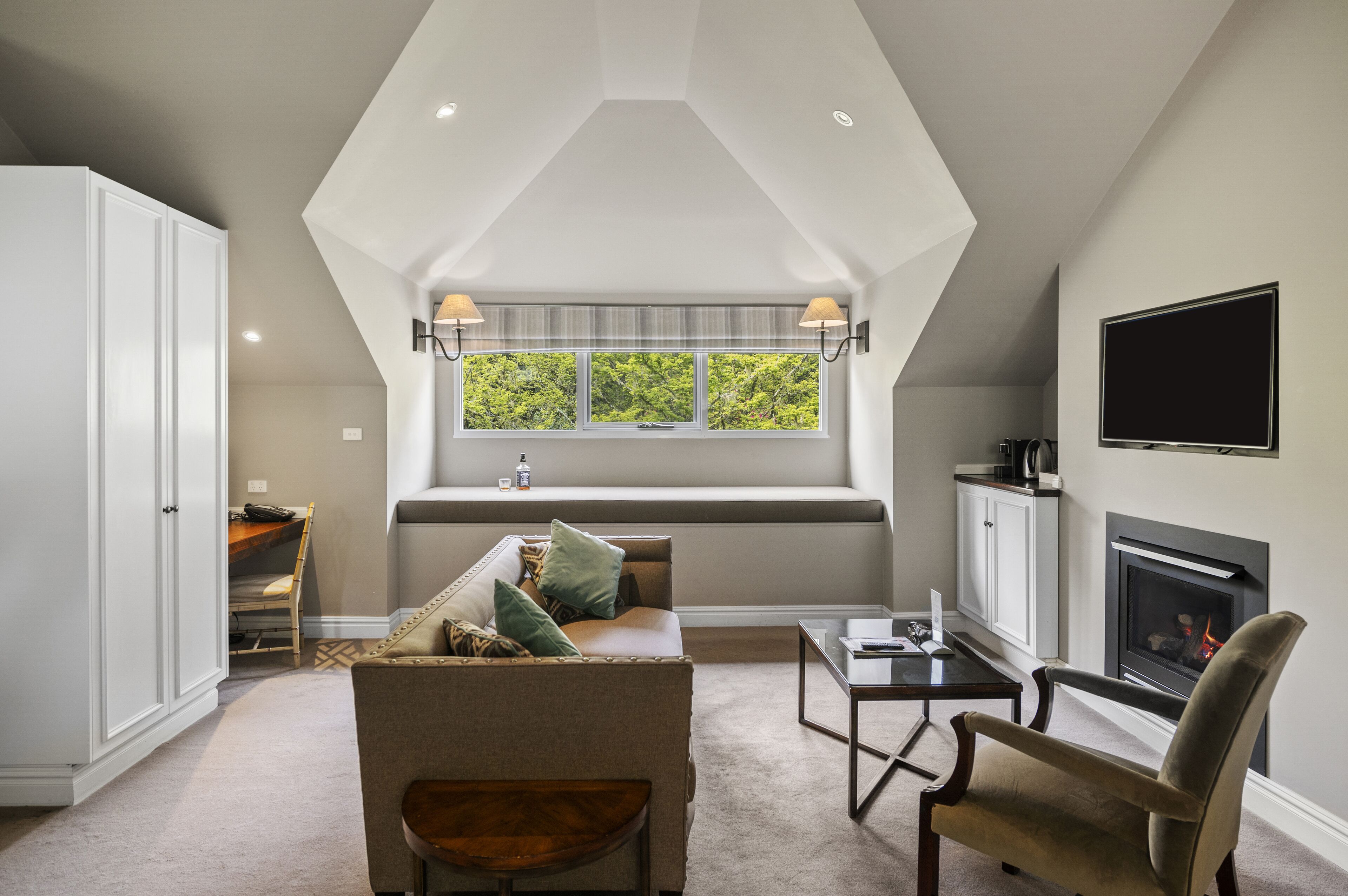 Ein Loft-Wohnzimmer mit hohen Decken, Kamin, großem Fenster mit Blick auf Bäume, einer Couch und einem an der Wand befestigten Fernseher.