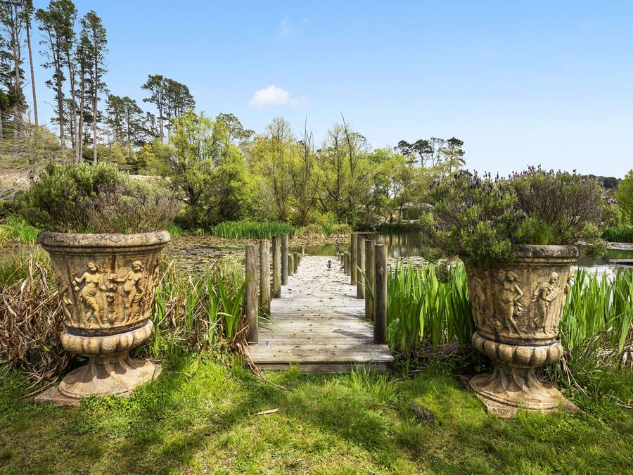 Un sentiero di legno conduce a uno stagno, affiancato da due grandi fioriere in pietra con sculture.