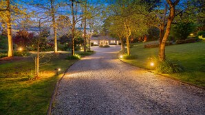 Een grindpad leidt naar een huis, omgeven door bomen, met een verzorgd gazon en een heldere lucht.
