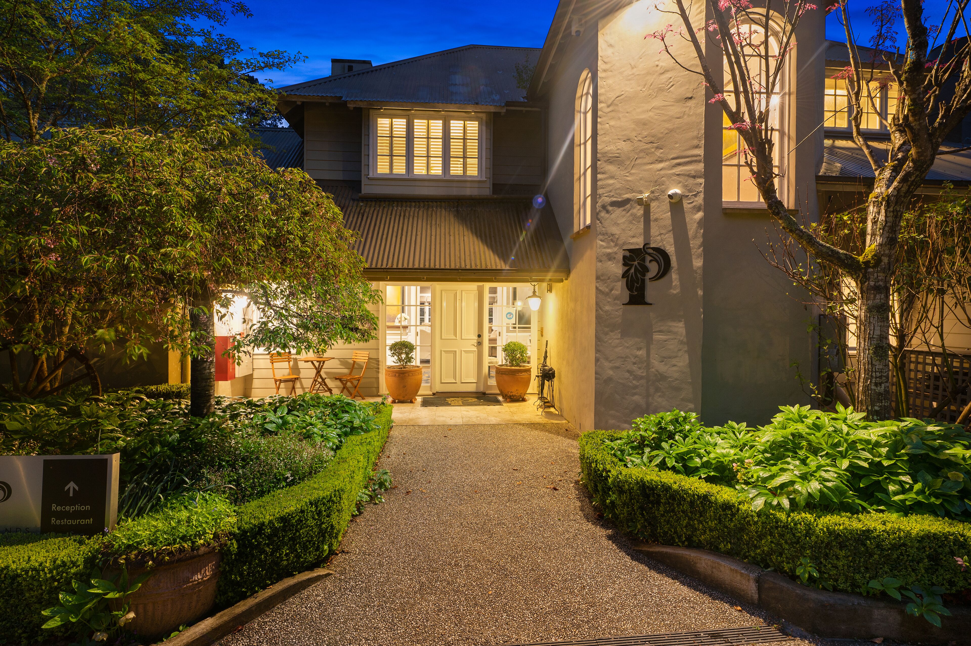 A well-lit reception area with a gravel path leading to the entrance, surrounded by neatly trimmed hedges and potted plants.