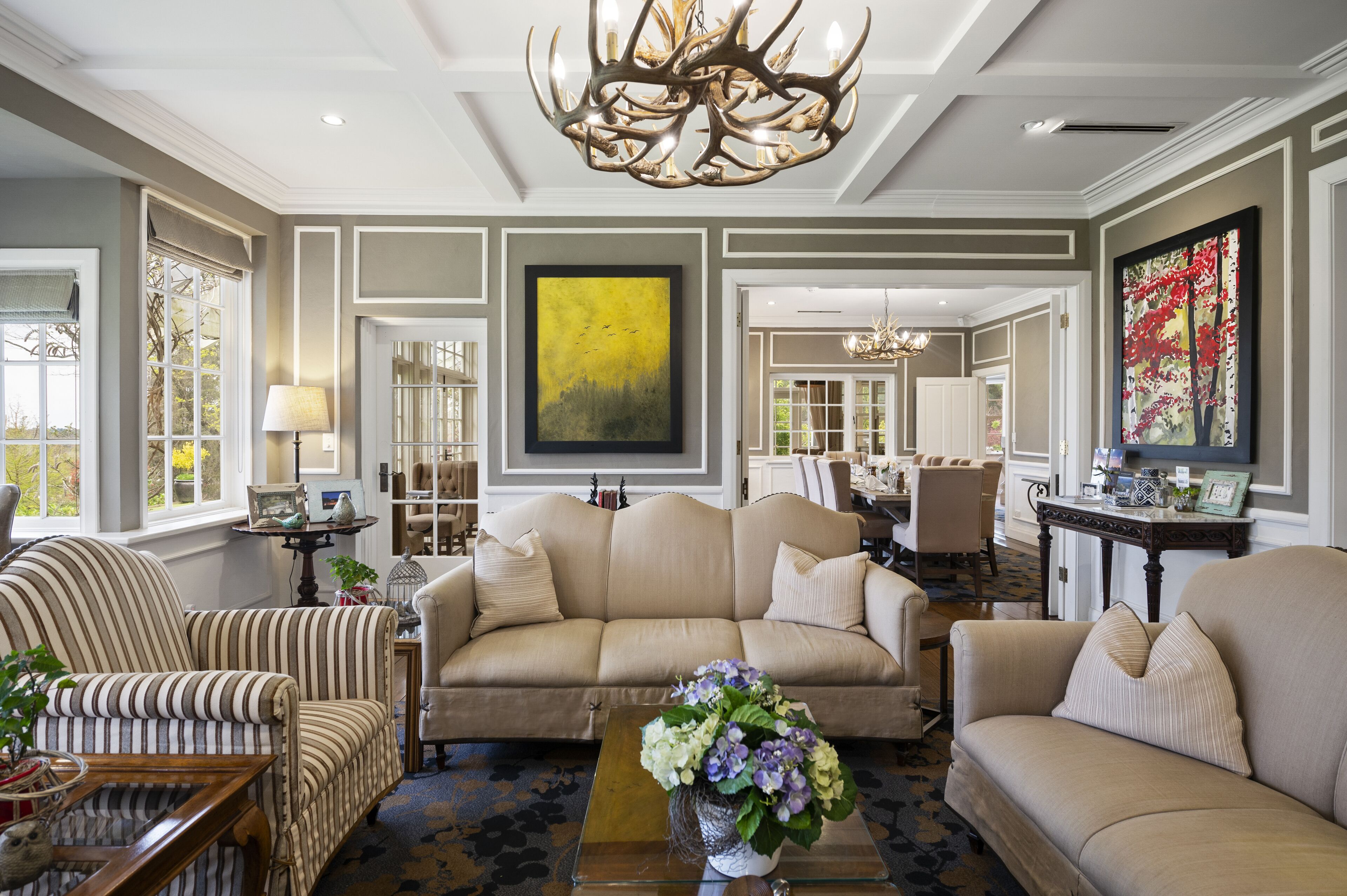 A spacious living room with a beige sofa, striped armchair, and a coffee table with a floral centerpiece. A large abstract painting and a chandelier are visible.
