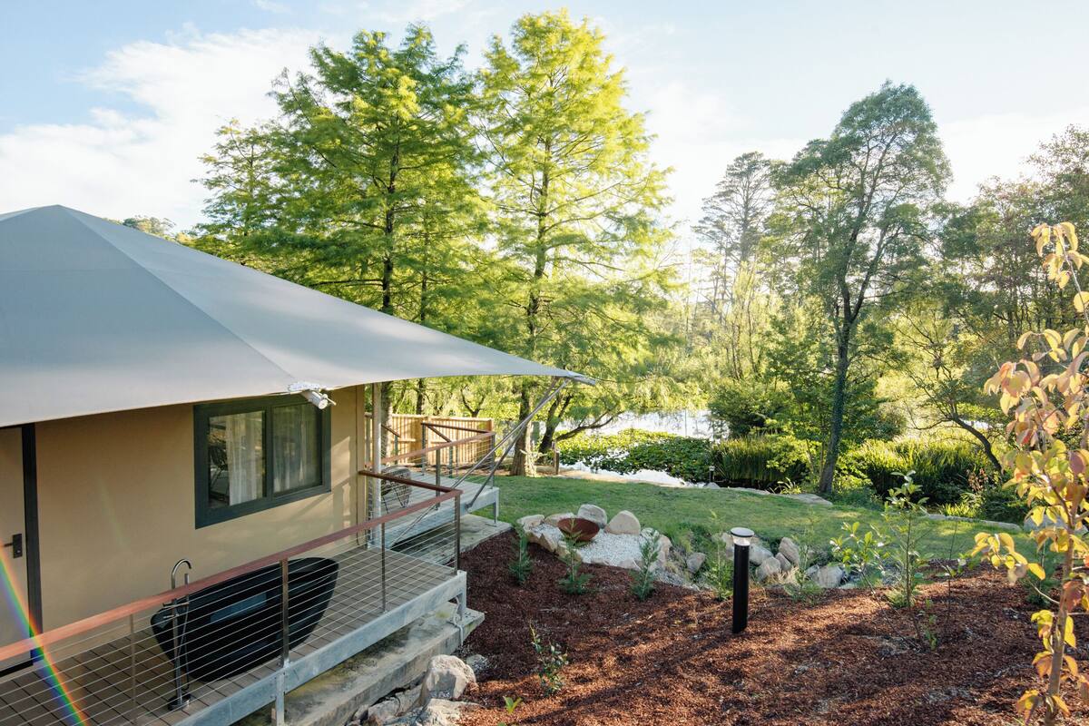 A modern RV with a covered porch, a small deck, and a landscaped garden with trees and a pond.