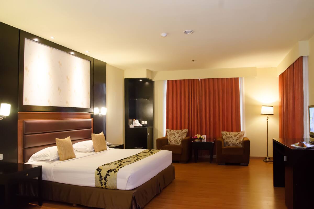 Habitación de hotel con una cama grande, dos sillones, un escritorio con televisor, un minibar y un ventanal con cortinas.