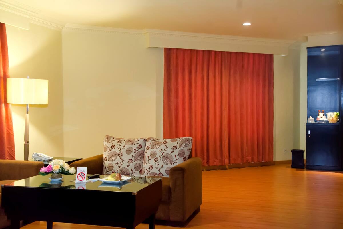 Habitación de hotel con sofá, una mesa con lámpara, una mesita con jarrón de flores y un ventanal con cortinas rojas.