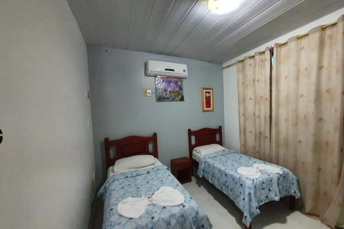 Un dormitorio con dos camas individuales, una mesita de noche, un equipo de aire acondicionado y una cortina.