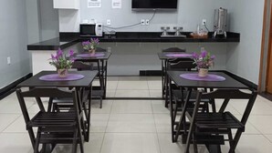 A modern kitchen with a flat-screen TV, a coffee machine, and a microwave. There are four tables with chairs, each with a purple placemat and a potted plant.